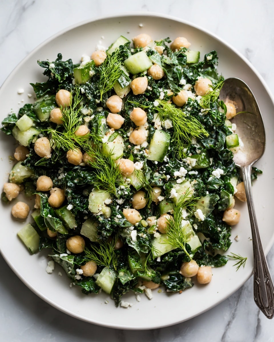 A white plate with a fresh kale and chickpea salad, showing chunky chopped dark green kale leaves mixed evenly with round pale beige chickpeas and light green cucumber cubes. Small white bits mix through the salad, likely cheese, with fresh bright green mint and dill sprigs placed on top as garnish. A silver spoon rests inside the plate on the right side, with the whole arrangement placed on a white marbled surface photo taken with an iphone --ar 4:5 --v 7
