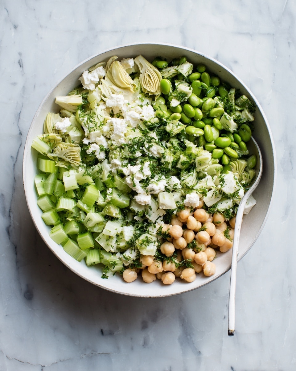 A white bowl filled with a vibrant salad that has layers of light beige chickpeas, bright green edamame beans, pale yellow artichoke hearts, chopped cucumber with a fresh light green color, small pieces of white feta cheese, and finely chopped green herbs sprinkled all over. A silver spoon is partly buried in the salad, resting on the right side of the bowl. The bowl sits on a white marbled surface with a soft texture. Photo taken with an iphone --ar 4:5 --v 7