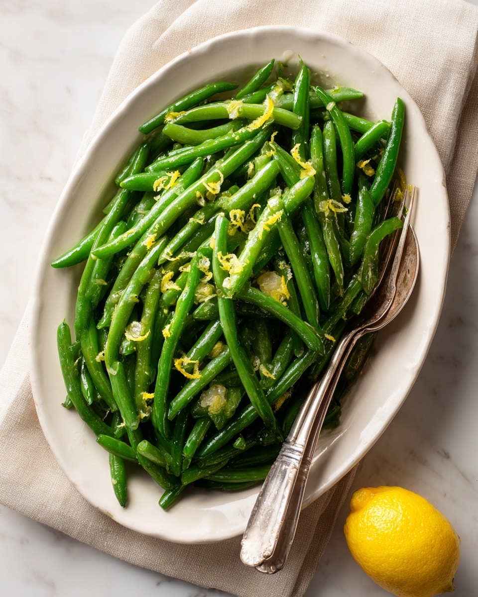 A white oval metal tray filled with a single layer of bright green cooked green beans that have a shiny texture, mixed with small yellow lemon zest pieces scattered evenly on top. A metal serving spoon and fork rest on the right edge of the tray. The tray is placed on a beige cloth on a white marbled surface. A whole yellow lemon is positioned near the bottom right corner of the image. Photo taken with an iphone --ar 4:5 --v 7