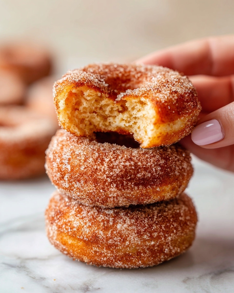The image shows three donuts on a metal cooling rack over a white marbled surface. The front donut is fully covered with a layer of coarse sugar and cinnamon, giving it a rough, grainy texture in light brown and white colors. Behind this donut, two more plain donuts have a smooth, slightly golden brown surface with no toppings. The donuts are round with a hole in the middle, and the cooling rack has black metal bars. Photo taken with an iphone --ar 4:5 --v 7