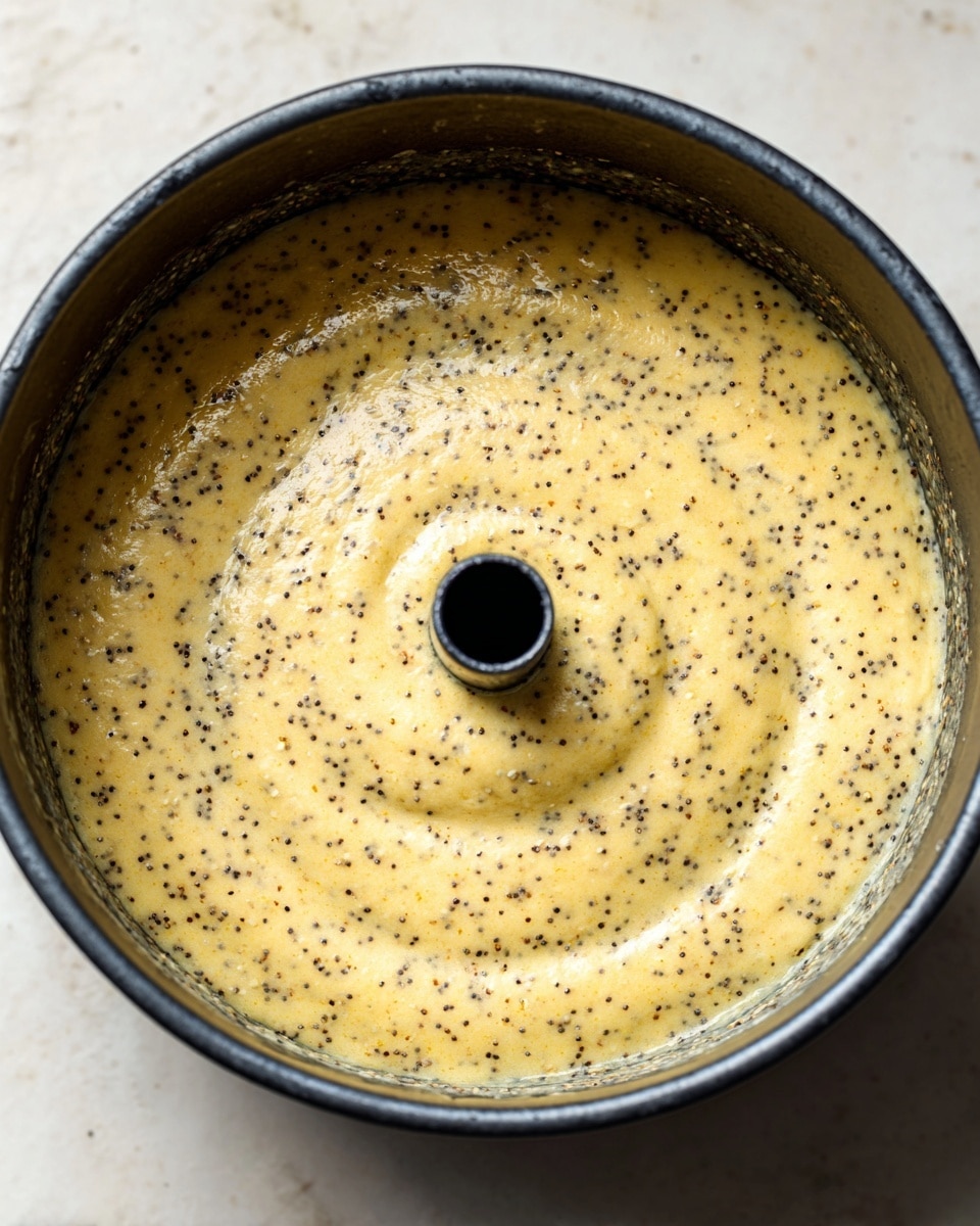 A close-up top view of unbaked poppy seed cake batter spread evenly in a dark metal bundt pan with a central tube. The batter is pale yellow with many tiny black poppy seeds mixed throughout, creating a speckled texture. The surface looks smooth but slightly uneven with soft folds. The pan is sitting on a white marbled surface. photo taken with an iphone --ar 4:5 --v 7
