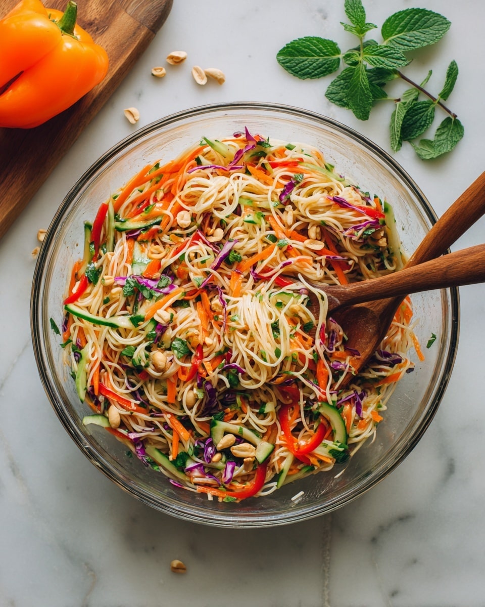 A large clear glass bowl filled with a bright and colorful noodle salad sits on a white marbled surface. The salad has three main layers: the bottom layer is thin, light beige rice noodles; the middle layer shows thin strips of orange carrots, red bell peppers, light green cucumber slices, and some purple cabbage; the top layer is sprinkled with chopped peanuts, green chopped herbs, and white sesame seeds. Two wooden spoons rest inside the bowl, one on each side. Around the bowl, there is a folded blue textured cloth on the left, a wooden board with fresh green mint leaves and a red bell pepper to the right, and an orange bell pepper in the upper right corner. Photo taken with an iphone --ar 4:5 --v 7