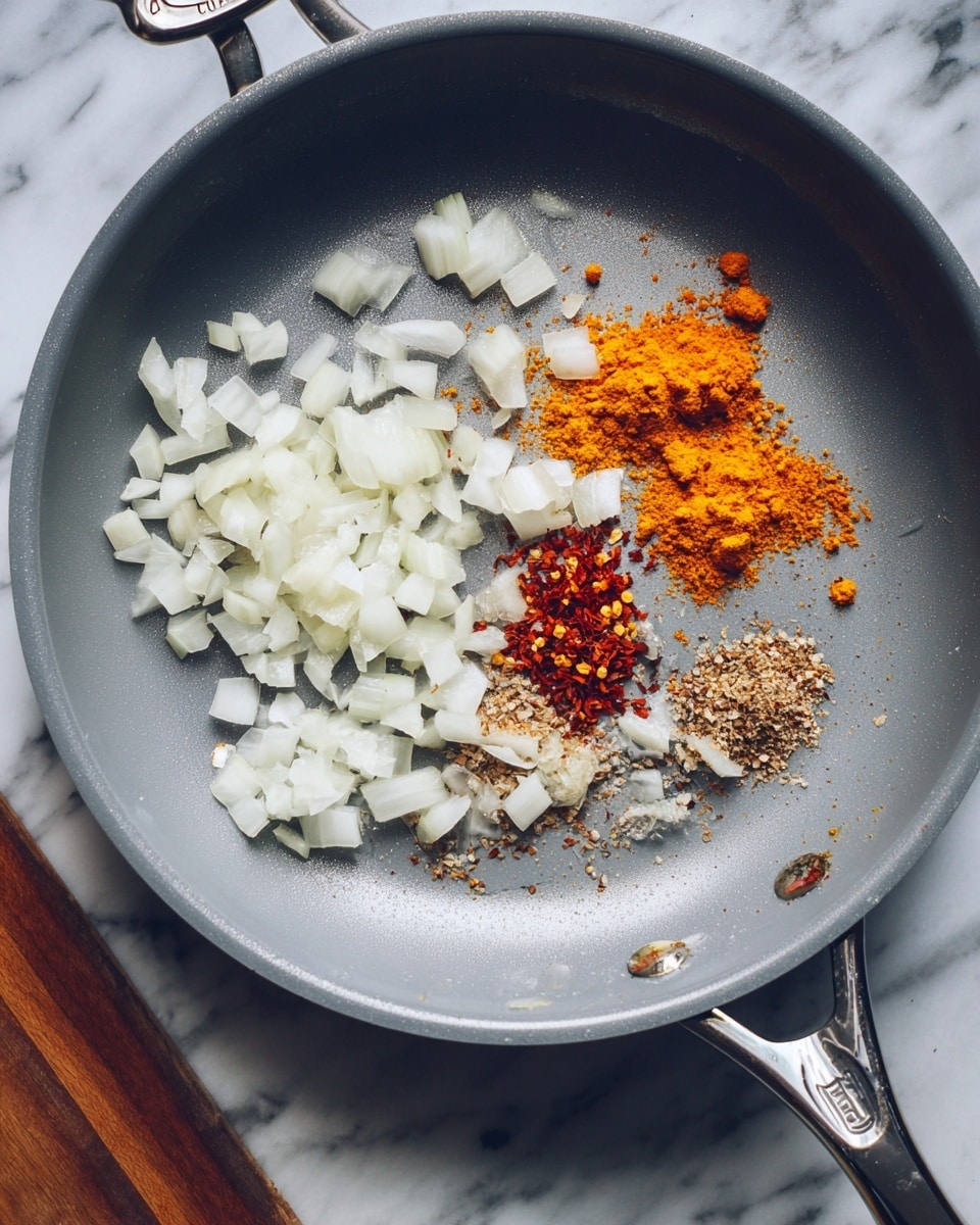 A grey frying pan with a shiny silver handle holds diced white onions spread out mostly on the left and right sides. Near the center are small piles of colorful spices: bright orange turmeric, reddish chili flakes, and light brown ground spices, arranged closely together. The pan sits on a white marbled surface with visible wood textures in the background. photo taken with an iphone --ar 4:5 --v 7