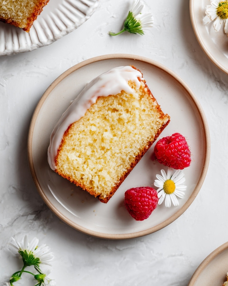 A single slice of yellow pound cake sits on a white plate with a light beige rim, the cake showing a moist, crumbly texture with a thin layer of white icing lightly covering the top edge. Two bright red raspberries rest next to the cake slice on the plate, with a small white daisy flower adding a delicate touch nearby. The plate is set on a white marbled surface, and parts of other lightly textured, white plates with raised edges are visible around the edges of the frame. Photo taken with an iphone --ar 4:5 --v 7