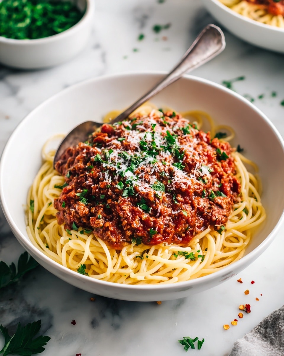 A white bowl filled with three layers: at the bottom, light yellow spaghetti noodles piled loosely; in the middle, a thick, chunky reddish-brown meat sauce with bits of vegetables; on top, small green parsley leaves and white grated cheese sprinkled evenly; a silver fork and spoon rest inside the bowl on the left side. The bowl sits on a white marbled surface with green parsley and red pepper flakes scattered around, with a smaller white bowl filled with green parsley in the blurred background. Photo taken with an iphone --ar 4:5 --v 7