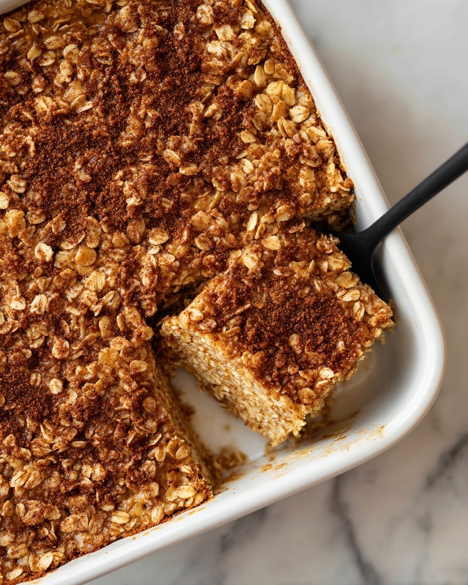 A white square baking dish holds a thick, textured oatmeal bake with a top layer full of chunky oats and soft, brown cinnamon pieces scattered through it. The oatmeal bake is golden brown with darker brown cinnamon powder sprinkled unevenly on top. A black spatula is placed inside the dish on the left side, slightly lifting a square piece from the corner, showing the same textured layer inside. The background is a white marbled surface. Photo taken with an iphone --ar 4:5 --v 7