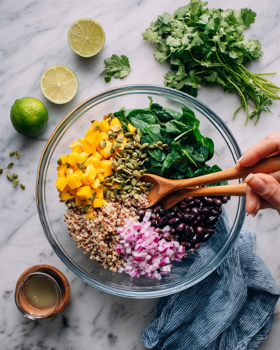 A clear glass bowl holds a colorful mix of fresh ingredients on a white marbled surface. At the bottom, there is a layer of dark green spinach leaves. On top of these leaves, there are small chopped bright yellow mango pieces on one side, finely chopped red onions with pink and white tones scattered across, and black beans in the upper right section. Mixed in are tan cooked quinoa grains and green pumpkin seeds, with some fresh green herbs like cilantro sprinkled around. A woman's hands hold two wooden spoons, stirring and mixing the vibrant ingredients inside the bowl. A bunch of fresh cilantro and halves of lime sit nearby, along with a small container of dressing. A blue-gray striped cloth is placed under the bowl's edge. Photo taken with an iphone --ar 4:5 --v 7