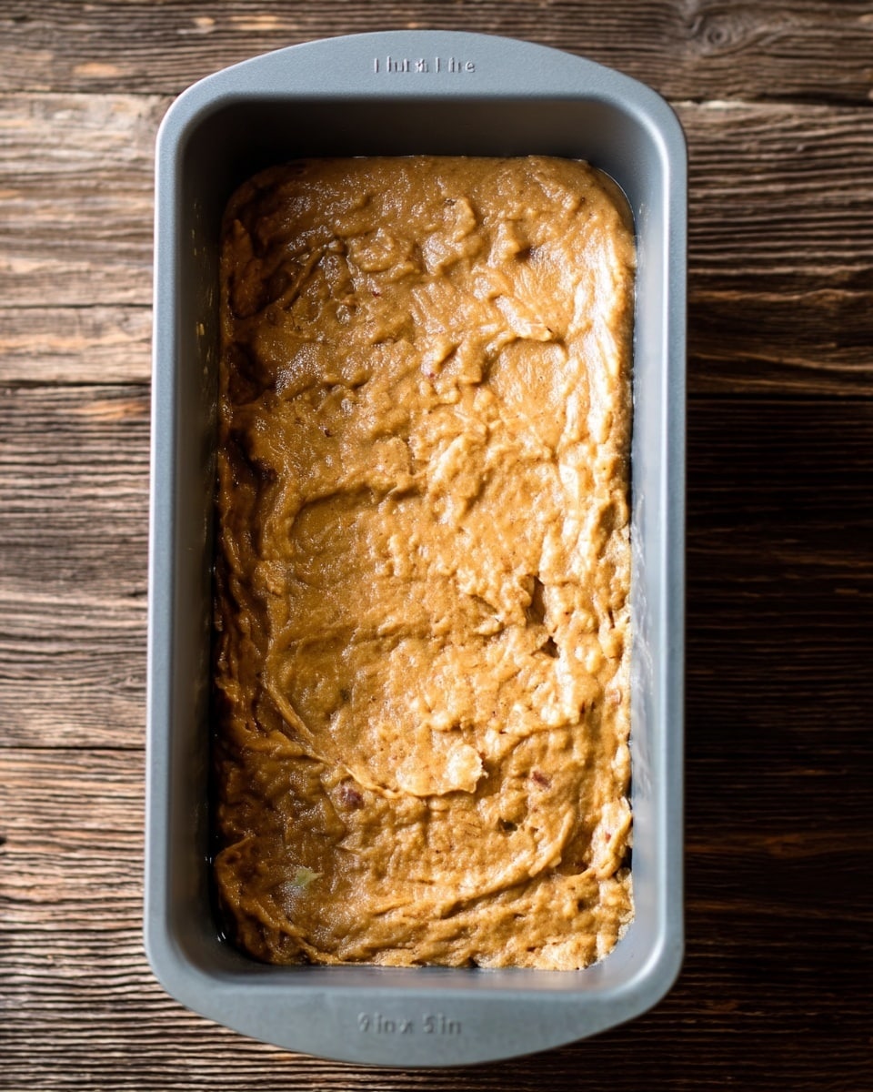 A gray baking pan filled with a thick, light brown batter that has a slightly uneven texture with small visible bits inside, spread evenly across the pan's surface. The pan sits on a wooden surface. The batter looks moist but firm, ready to be baked. photo taken with an iphone --ar 4:5 --v 7