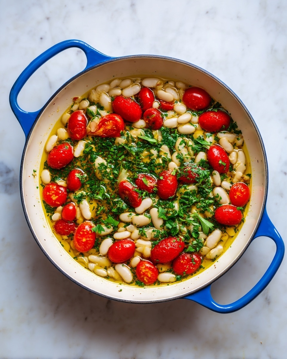 The image shows a shallow white pan with blue handles filled with a dish made of three main layers. The bottom layer is light golden oil with some herbs floating on it, giving a shiny texture. The middle layer has many small white beans, scattered evenly across the pan. On top of the beans are bright red cherry tomatoes, some whole and some sliced, adding color contrast. Sprinkled throughout are pieces of fresh green parsley, adding vibrancy and freshness. The pan is placed on a white marbled surface. Photo taken with an iphone --ar 4:5 --v 7
