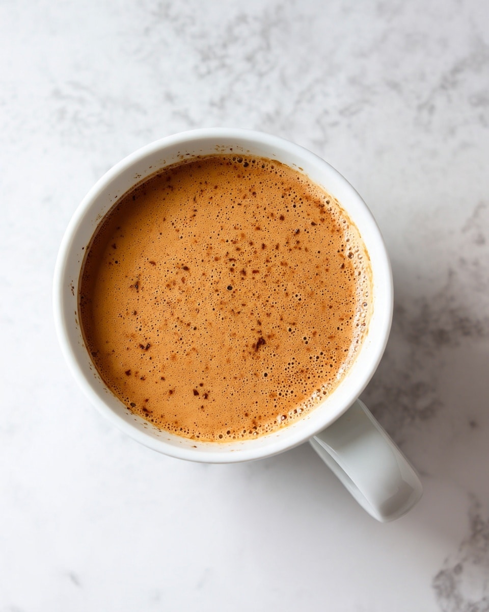 A white cup filled with a smooth light brown drink with tiny darker brown specks floating on the surface, placed on a white marbled texture background, top view showing the round shape of the cup and its handle extending to the right photo taken with an iphone --ar 4:5 --v 7