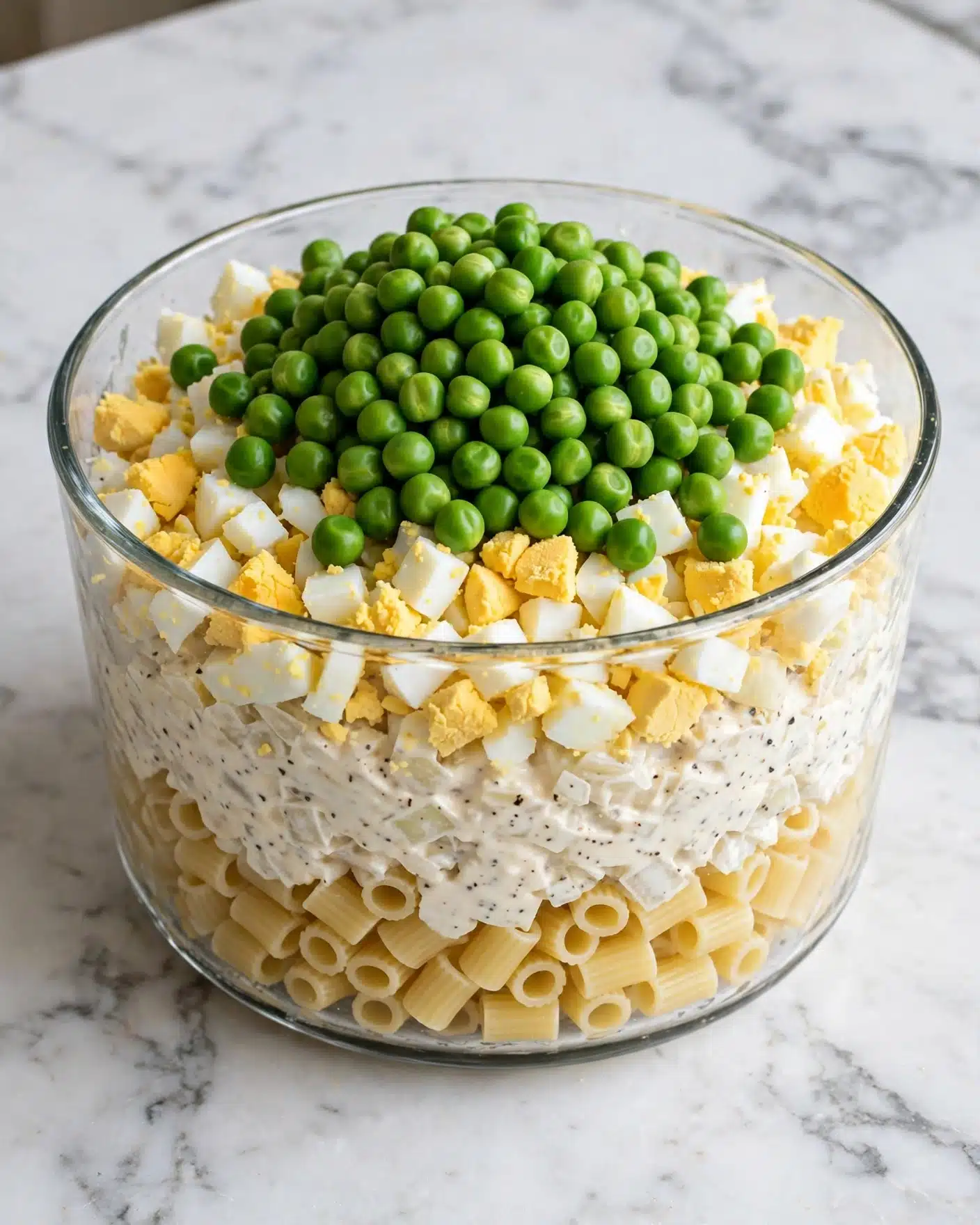 Tuna Macaroni Salad Recipe 5 A clear glass bowl holds a layered pasta salad on a white marbled surface. At the bottom, short tubular pasta pieces form a pale yellow base. On top of the pasta, there is a mix of small white chopped onions and creamy white mayonnaise with visible black pepper specks. Above this is a layer of chopped hard-boiled eggs, showing bright yellow yolks mixed with white parts in small cubes. The top layer is a mound of vibrant green peas sitting in the center, bright and fresh-looking. Photo taken with an iphone --ar 4:5 --v 7