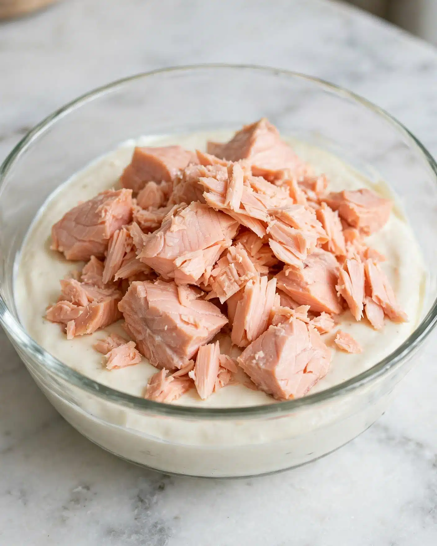 Tuna Macaroni Salad Recipe 4 A clear glass bowl sits on a white marbled surface, filled with a creamy white sauce spread evenly at the bottom. On top of this sauce, there is a generous pile of light pink, flaky salmon pieces, some chunks larger and others smaller, with a soft texture. The salmon looks tender and fresh, contrasting nicely with the smooth sauce beneath. photo taken with an iphone --ar 4:5 --v 7
